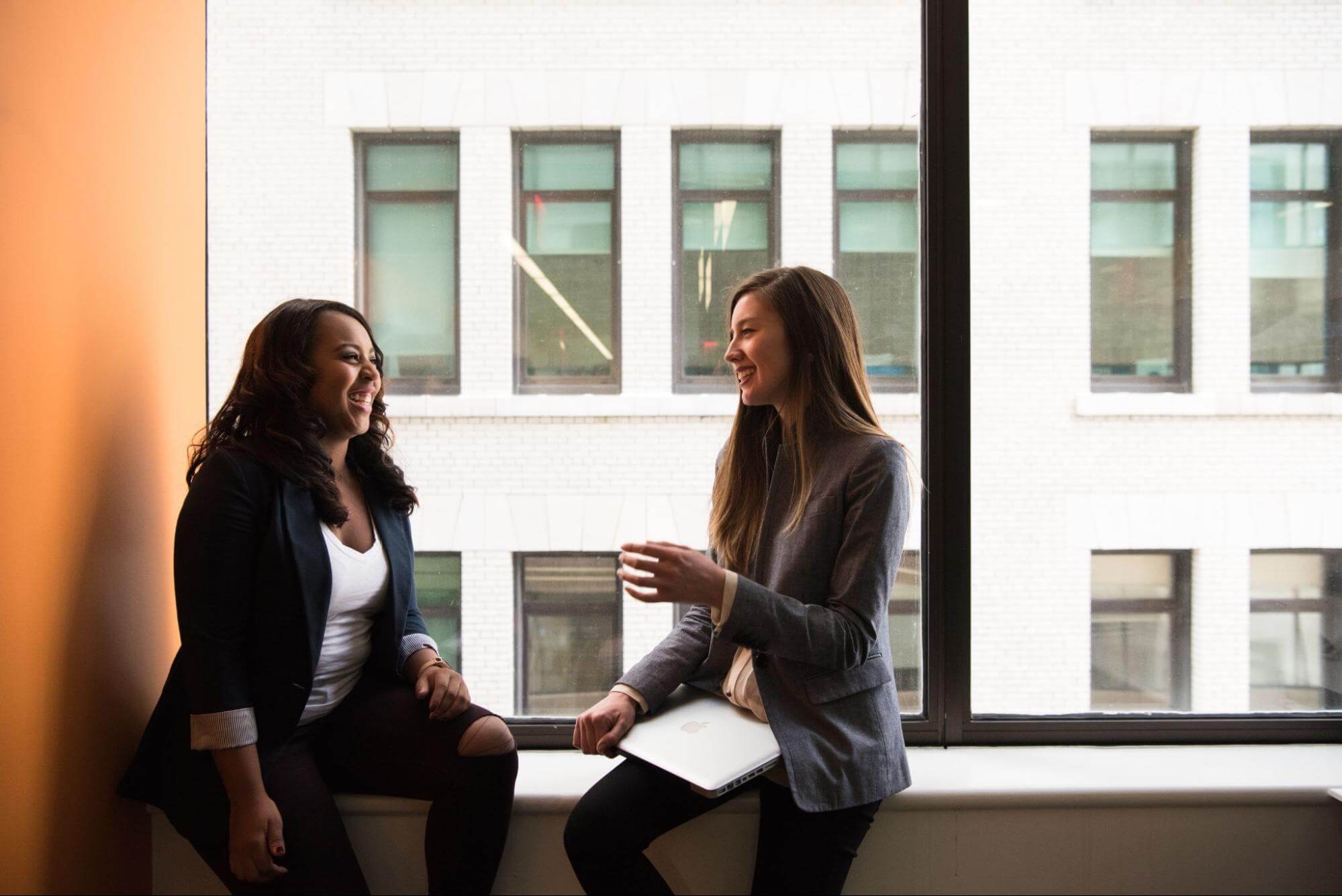 Two women smiling, chatting at window in office